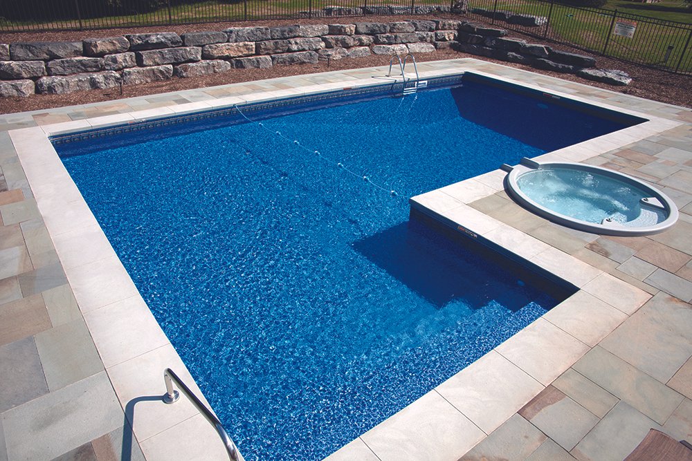 Swimming pool with a smaller hot tub attached, surrounded by a stone patio and a low stone wall, with a metal fence in the background.