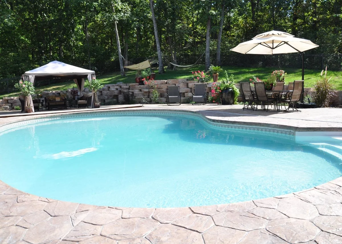An outdoor swimming pool surrounded by a stone deck, with patio furniture, a canopy, umbrellas, a flower garden, and trees in the background.