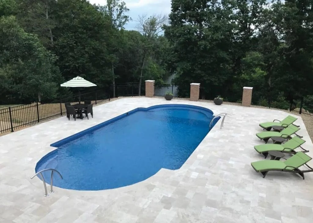 A backyard pool area with green lounge chairs, a small round table with four chairs, and a white umbrella, surrounded by trees.