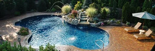 Backyard with a freeform swimming pool, surrounded by a brick patio, garden plants, and a large patio umbrella with lounge chairs.