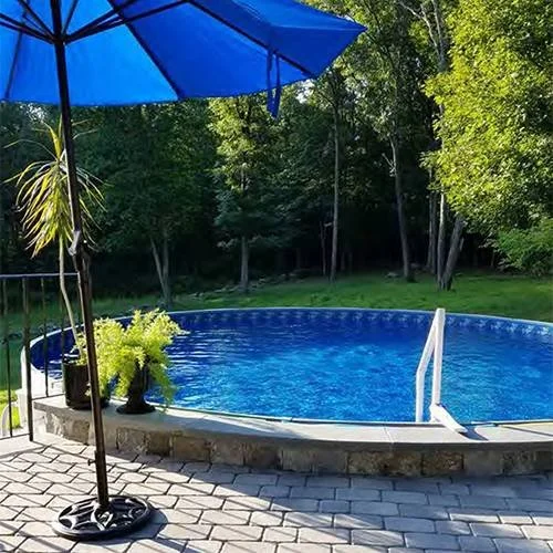 Round above-ground swimming pool with a blue cover, located on a stone patio, surrounded by green trees, with a blue umbrella, potted plants, and a white pool ladder.