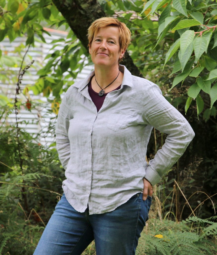 A woman with short reddish hair stands outdoors among green trees and plants, wearing a light-colored, rolled-up sleeve button-up shirt and jeans, smiling softly at the camera.