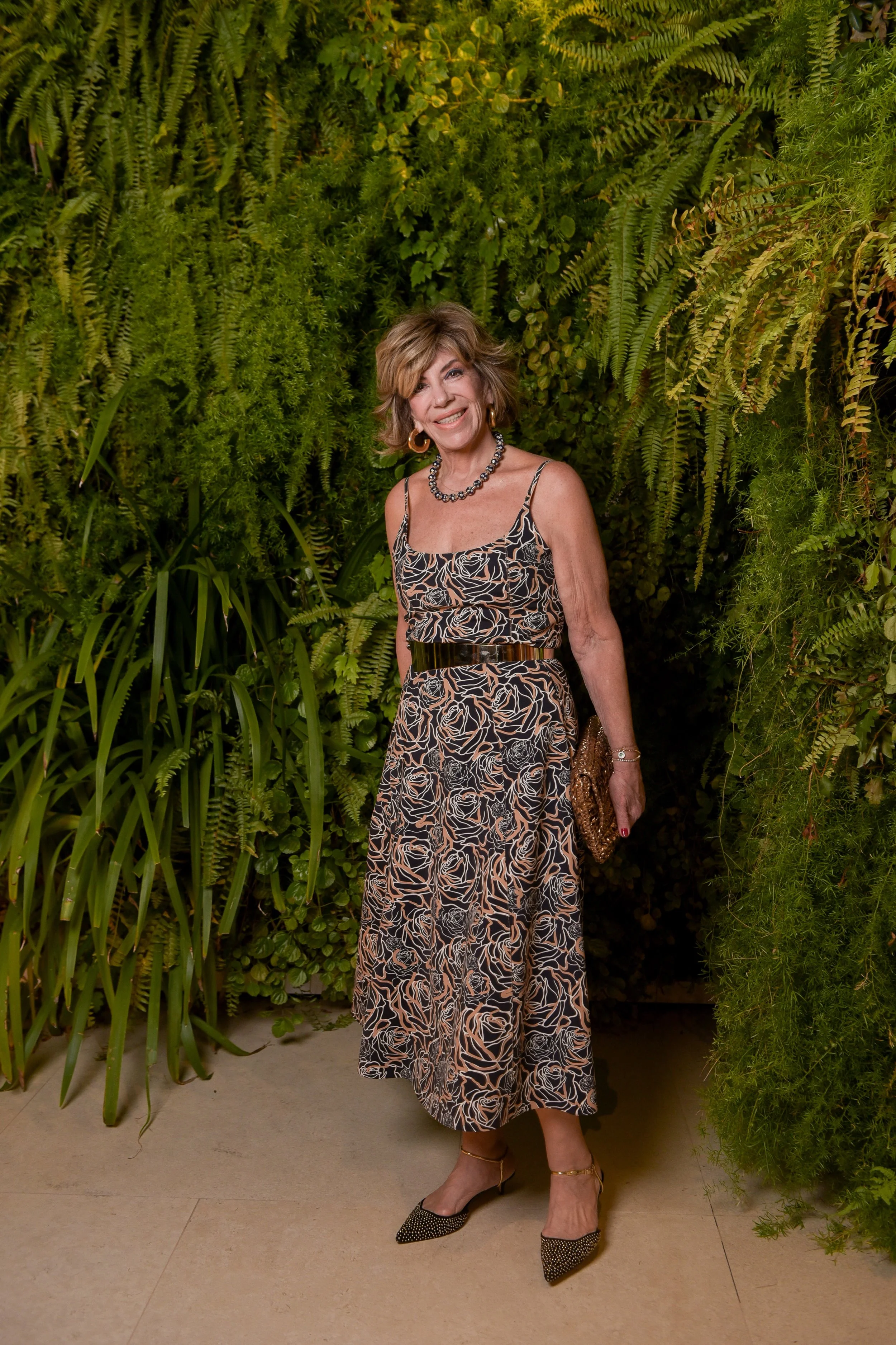 Mulher idosa com cabelo curto e castanho claro, usando vestido sem mangas com estampa de flores em tons de preto, bege e branco, brincos grandes, colar de pérolas e pulseira, segurando uma bolsa de mão com lantejoulas douradas. Ela está sorrindo e po