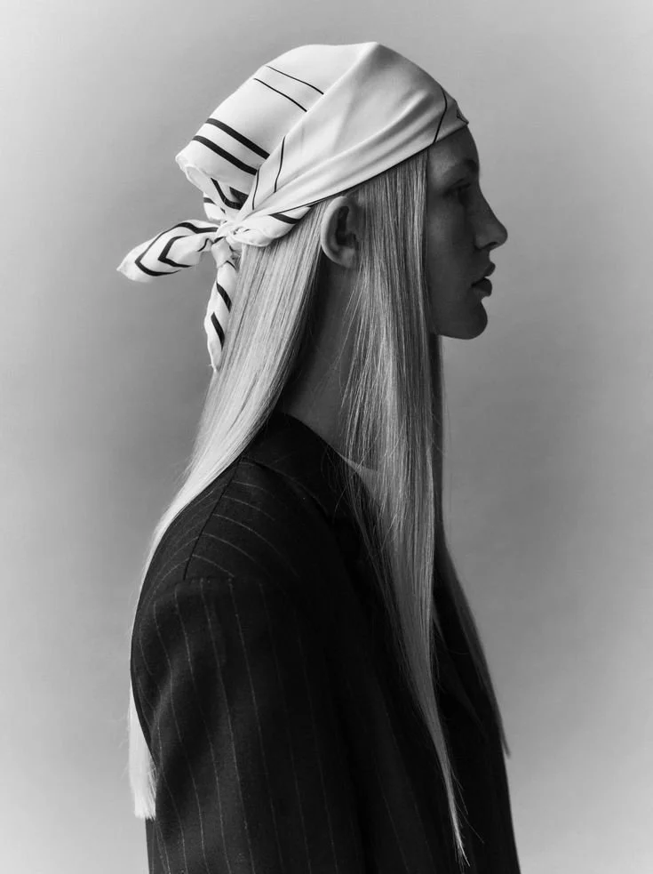 Mulher com cabelo liso e comprido, usando bandana branca com linhas pretas, vista de perfil, de roupa escura