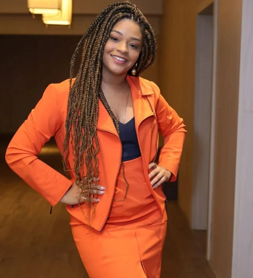 Mulher sorridente com cabelos enrolados, vestindo roupa laranja e blusa azul, em ambiente interno.