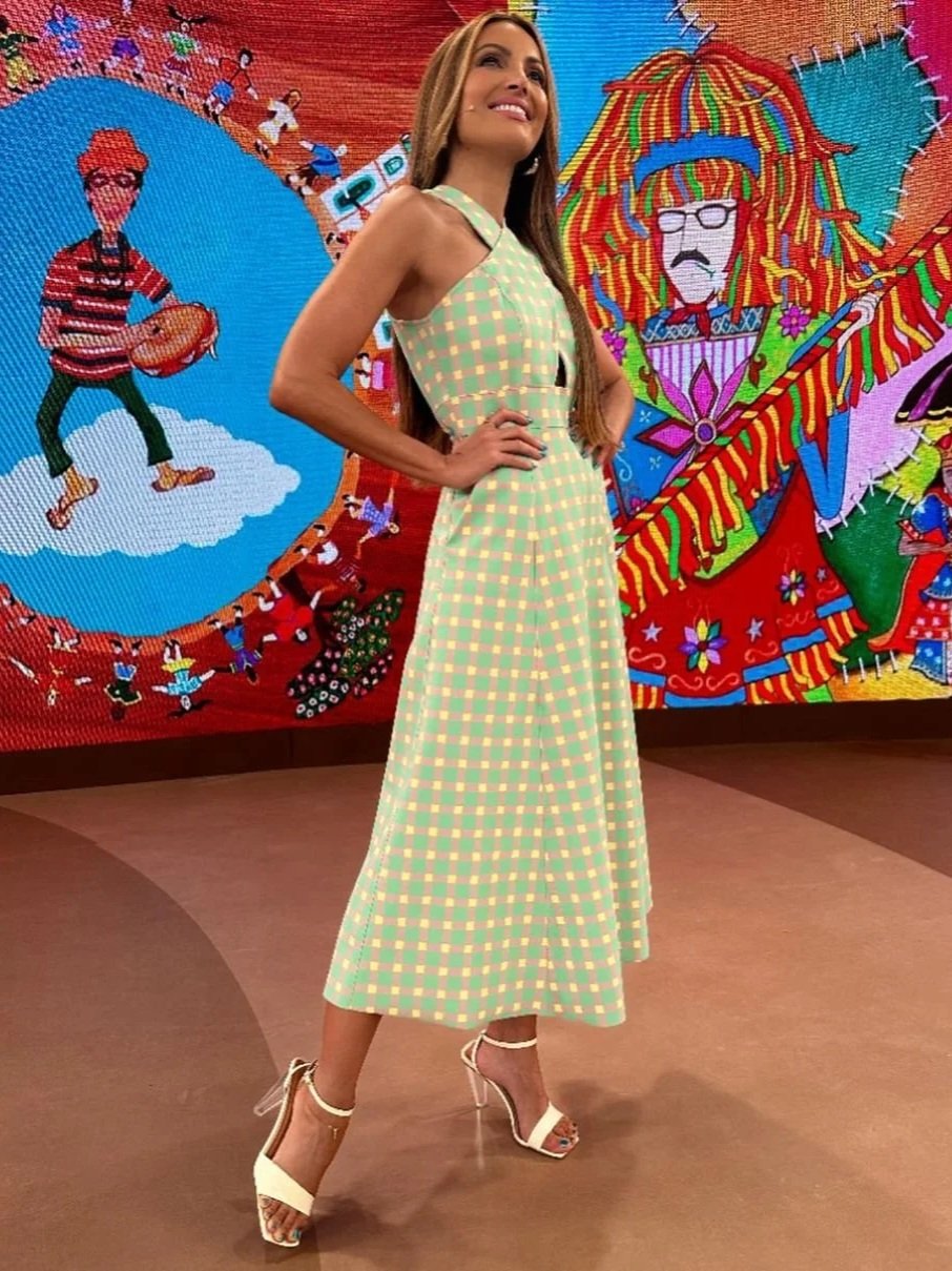 Mulher com vestido longo de estampa xadrez amarelo e verde e sapatos brancos de salto, posando com as mãos na cintura diante de um mural colorido de arte