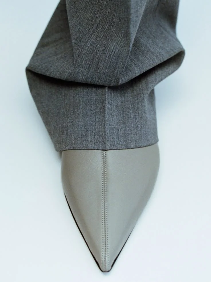 Close-up de um sapato de salto alto em tom bege, com o cabedal de textura suave, combinado com uma roupa cinza escura.