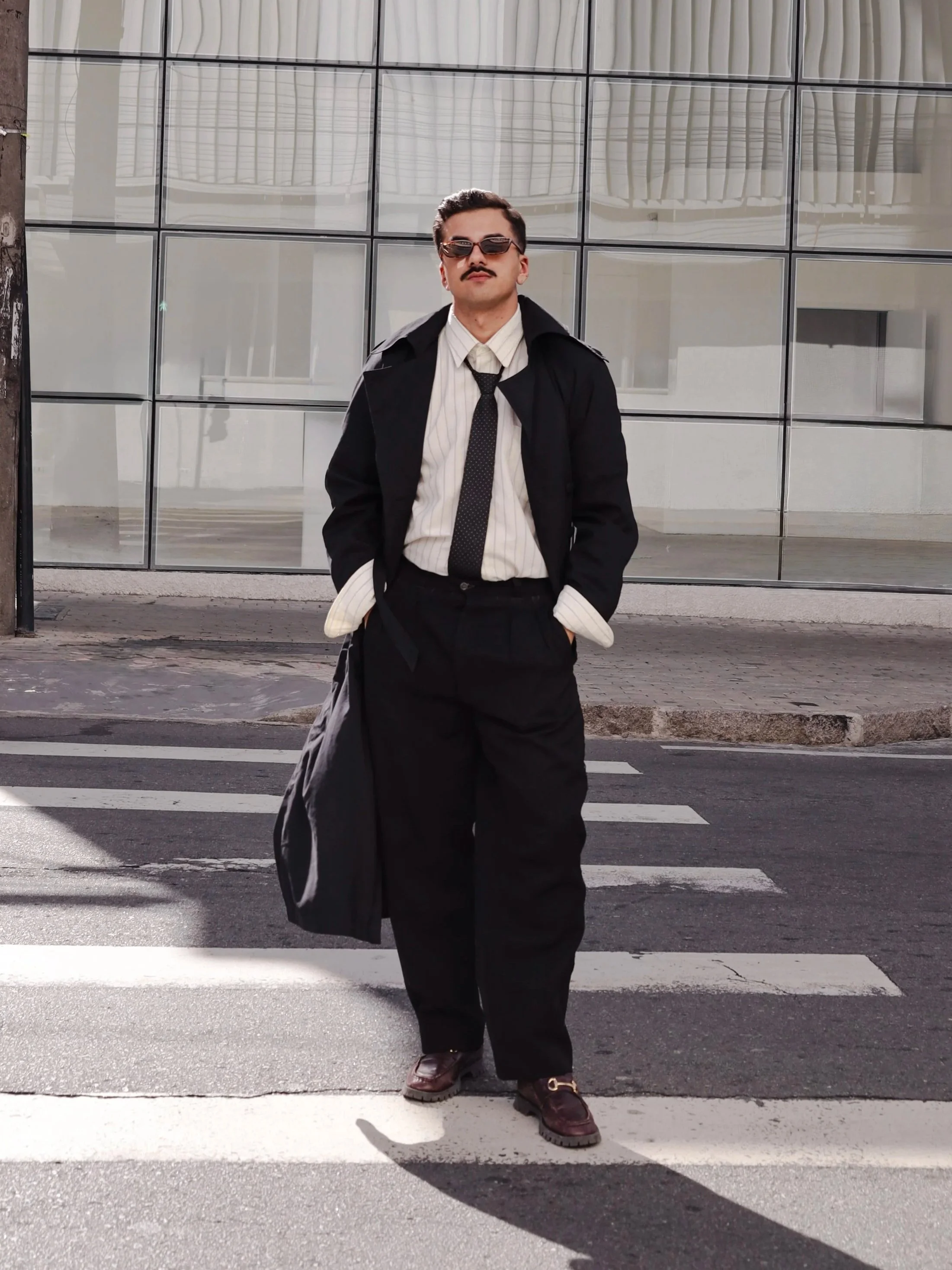 Homem com estilo vintage, camisete listrada, blazer preto, cabelo penteado, mustache, óculos escuros, calça preta, sapato marrom, segurando um sobretudo preto, posando na rua em frente a uma parede de vidro.