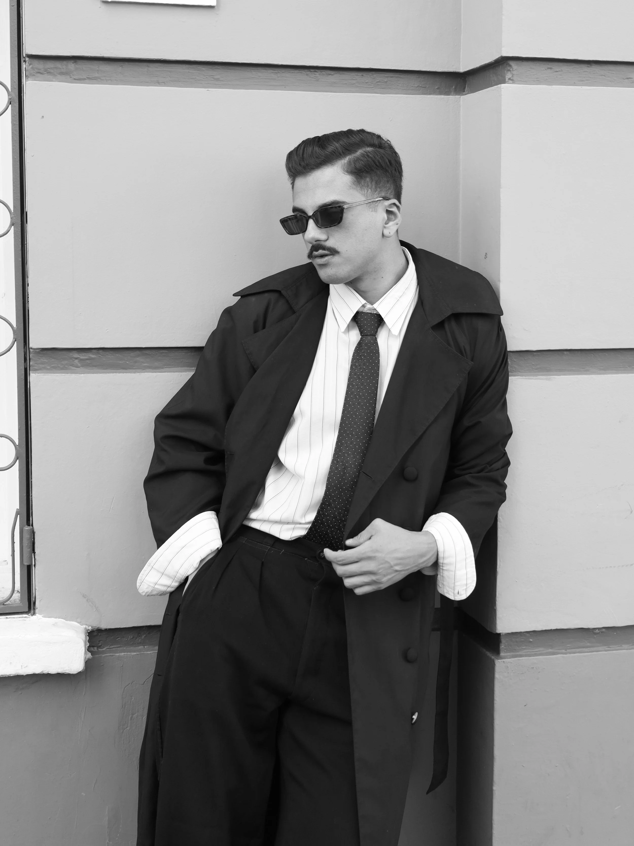 Homem com cabelo penteado, óculos escuros, bigode, usando terno, gravata e casaco longo, posando contra uma parede, na foto em preto e branco.