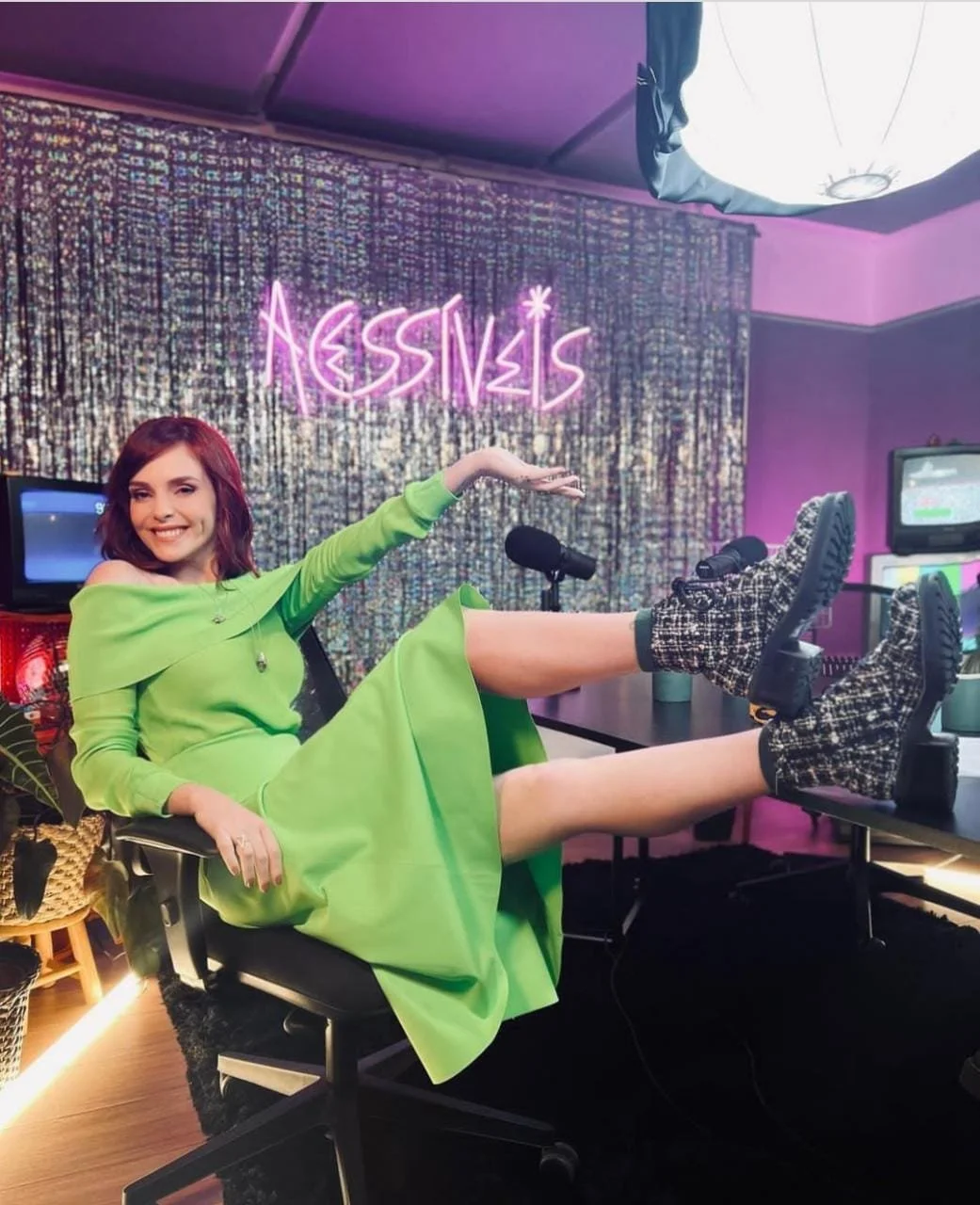 Mulher com cabelo vermelho, sorrindo, usando vestido verde, sentado em cadeira de escritório com pernas erguidas na mesa, usando botas de abuse com estampa de tweed, em um estúdio decorado com cortina brilhante, iluminação rosa, código neon 'AESSIS' 