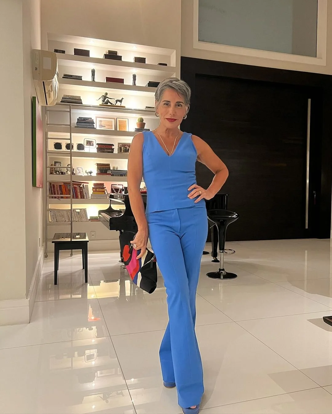 Mulher idosa de cabelo grisalho usando roupa azul, posando em sala de estar moderna com prateleiras de livros e decoração.