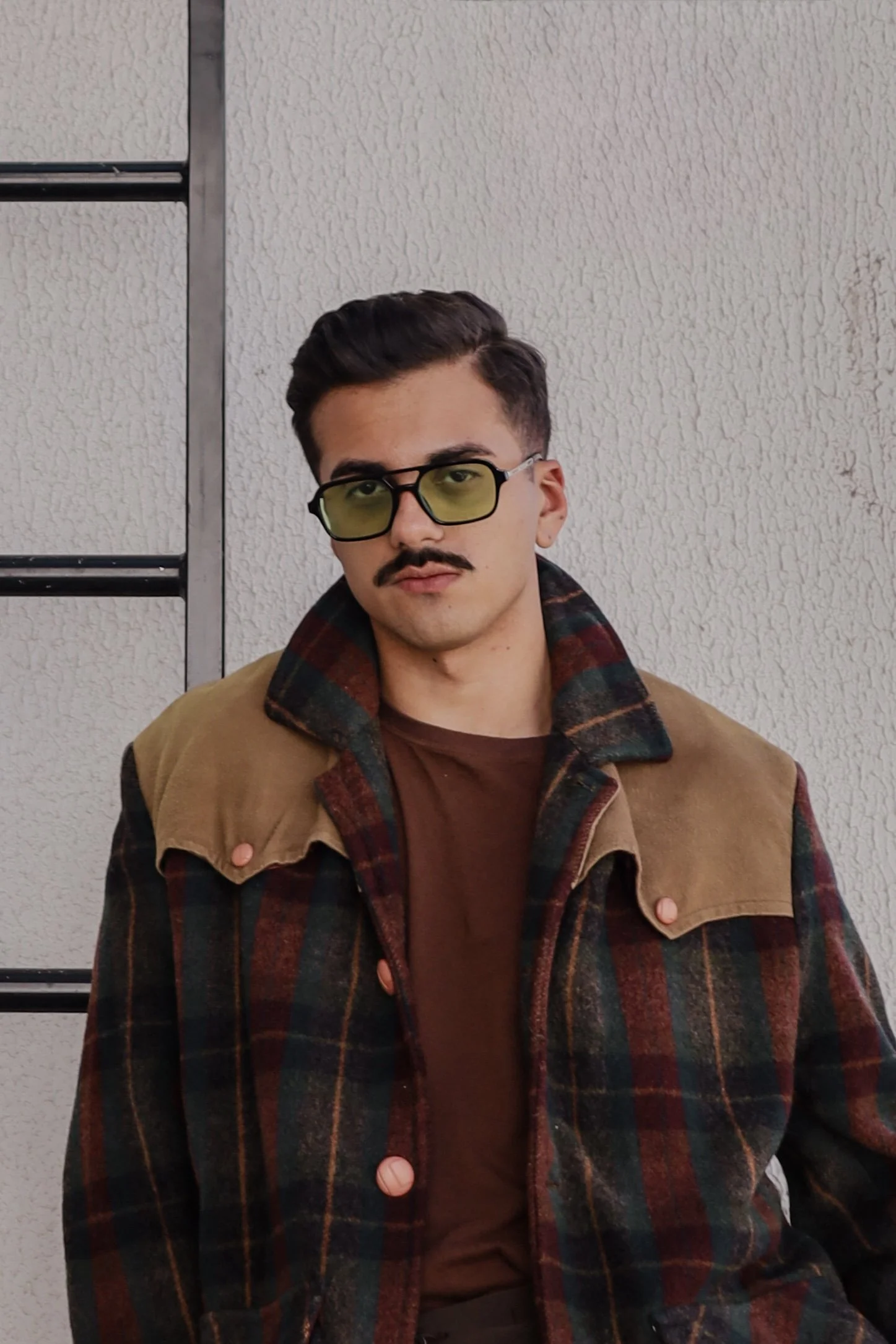 Homem com cabelo escuro, bigode e óculos de lente amarela, usando jaqueta xadrez marrom e camiseta marrom, posando contra parede de textura rugosa na cor branca.
