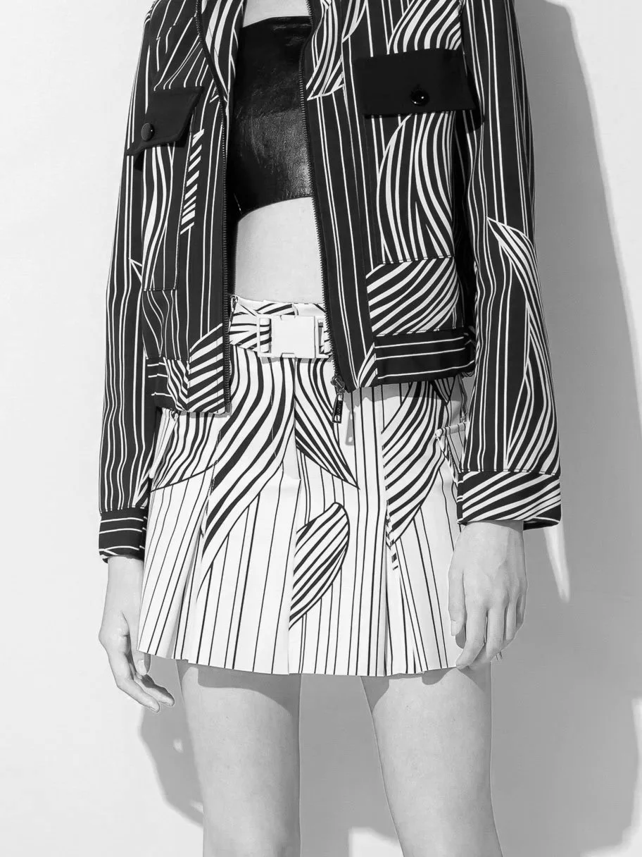 Pessoa de roupas de estilo moderno, usando uma jaqueta de listras preto e branco, uma saia de listras também preta e branca, com cinto branco, em fundo branco.