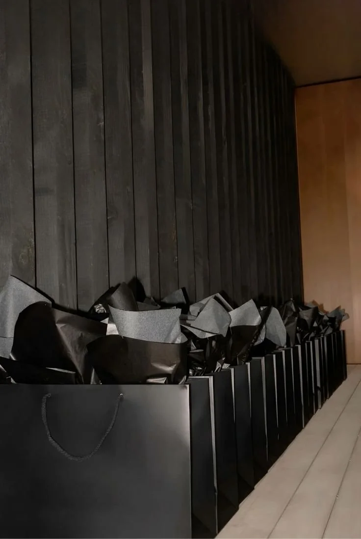 Vários sacos pretos e cinzas em bolsas de papel preto empilhados ao lado de uma parede de madeira escura.