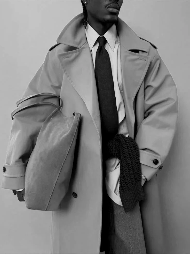 Homem vestido com trench coat, camisa branca, gravata, segurando uma bolsa grande, com uma blusa de manga comprida ao lado, em preto e branco.