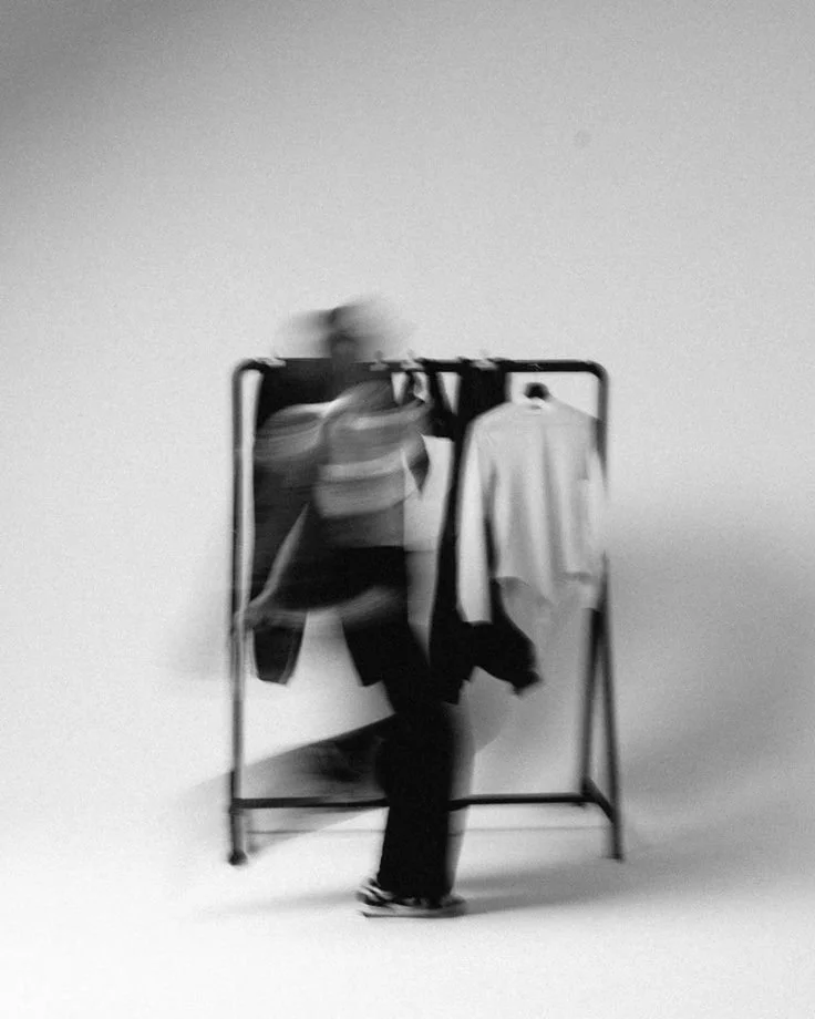 Pessoa em movimento na frente de um cabideiro com roupas, em fundo branco, foto em preto e branco com desfoque.