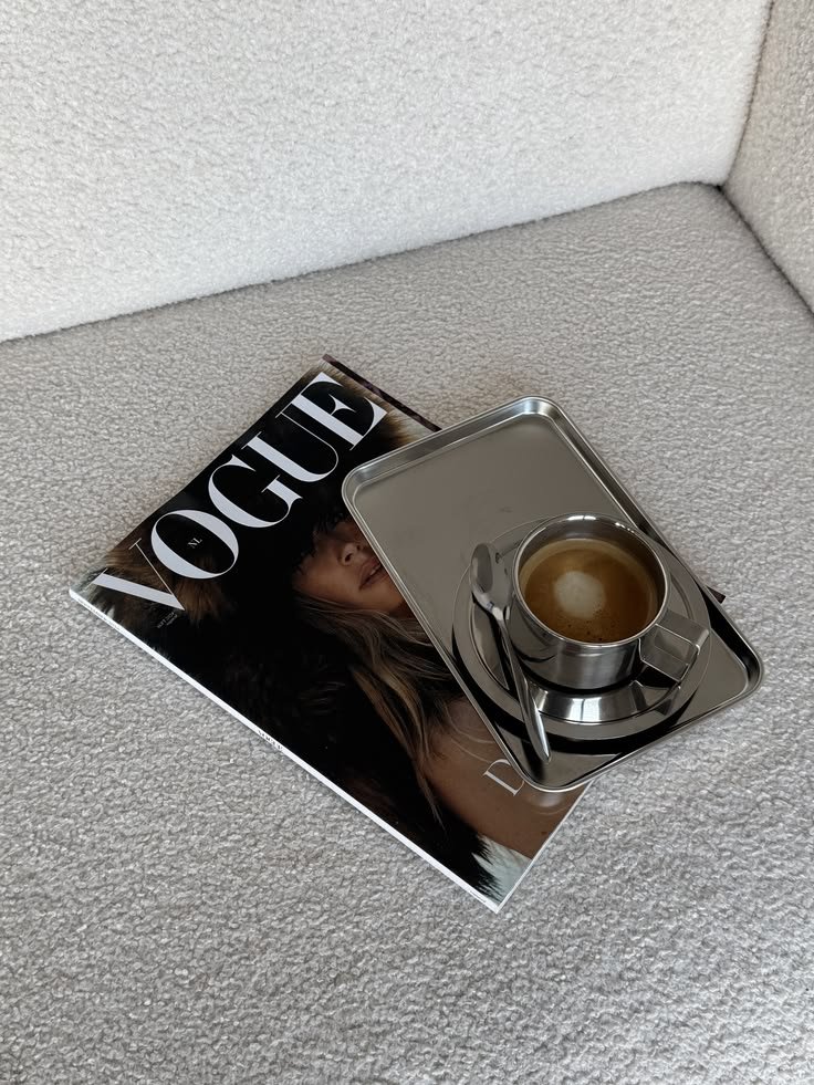 Revista Vogue brasileira, com uma xícara de café em uma bandeja de metal sobre ela, em um sofá claro.