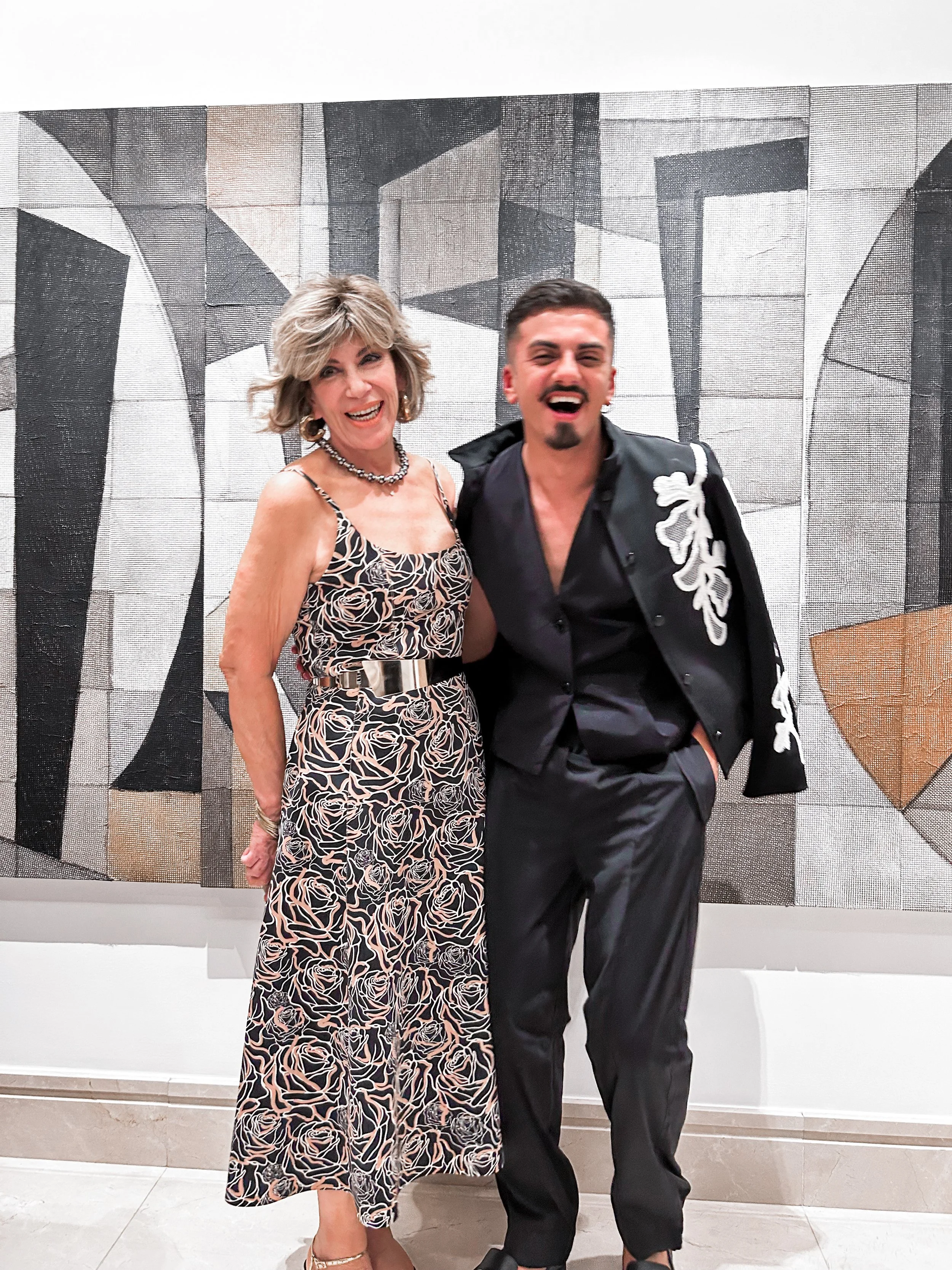 Mulher e homem sorrindo, posando em frente a uma parede com arte moderna, ela usando vestido longo com estampa de rosas e ele de terno preto com bordado branco, ambos felizes.