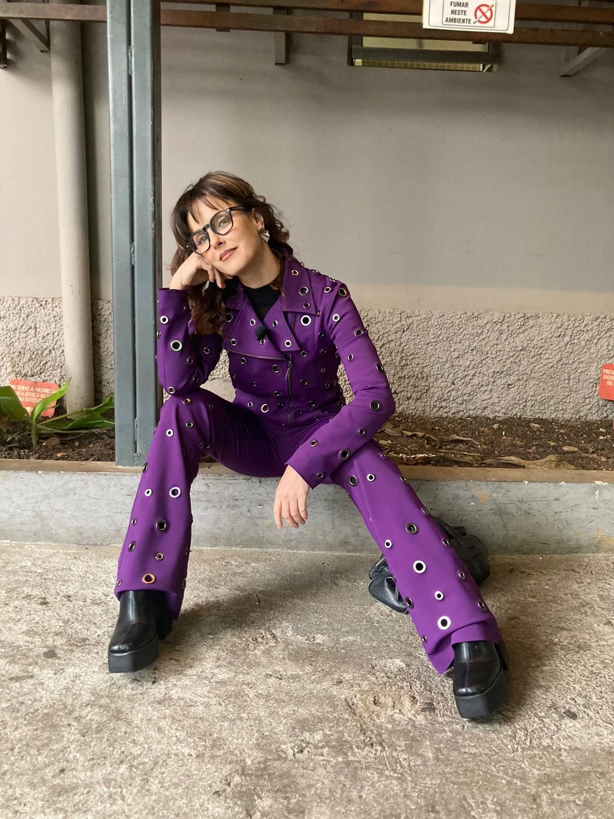 Mulher de óculos com cabelo cacheado, usando um conjunto roxo com olhos em uma calça e jaqueta de couro, sentada em um parapeito de cimento, com uma expressão pensativa.