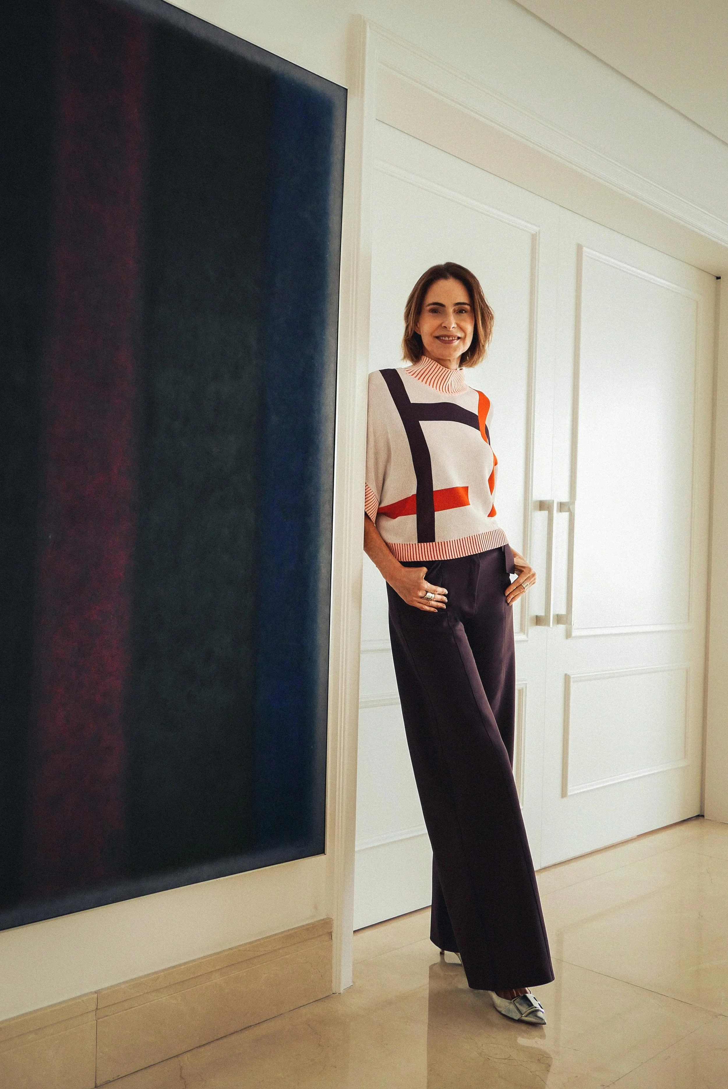 Mulher sorridente usando blusa branca com detalhes vermelhos, pretas e laranja, calça preta, posicionada ao lado de uma parede branca e um quadro de arte, no interior de uma residência ou galeria.