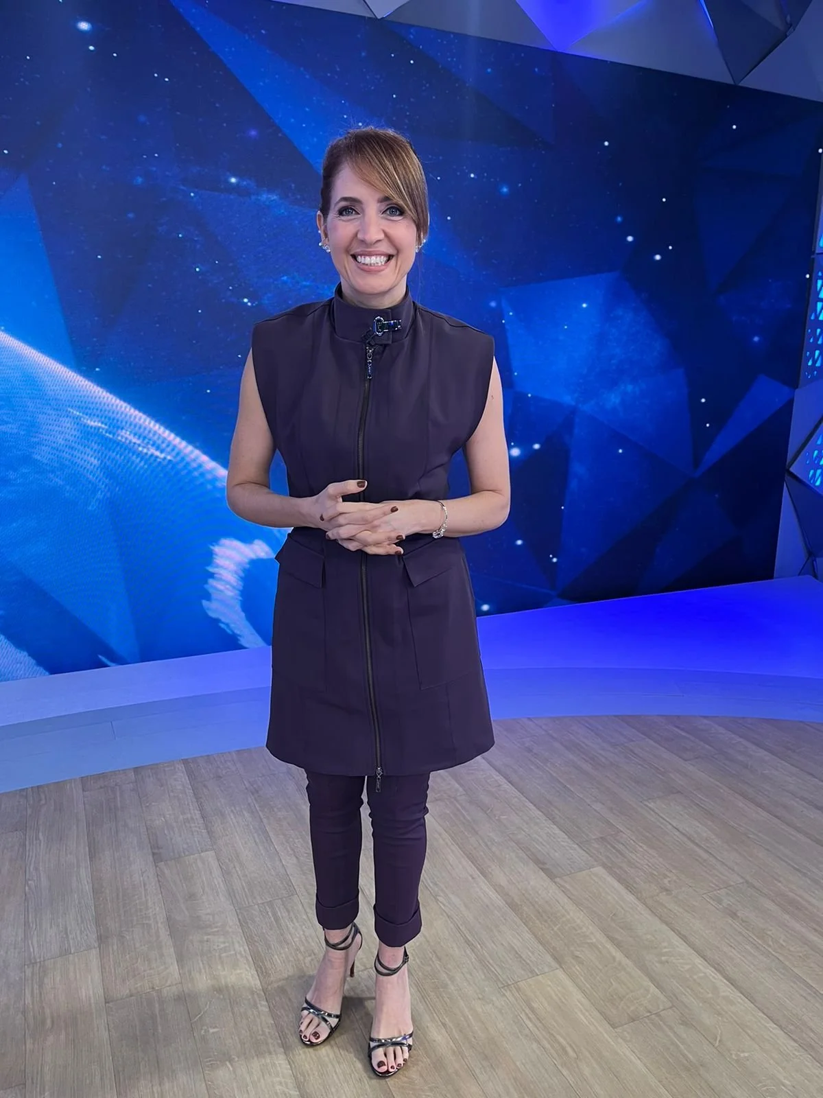 Mulher sorridente com vestido preto em estúdio de TV com fundo de espaço estrelado.