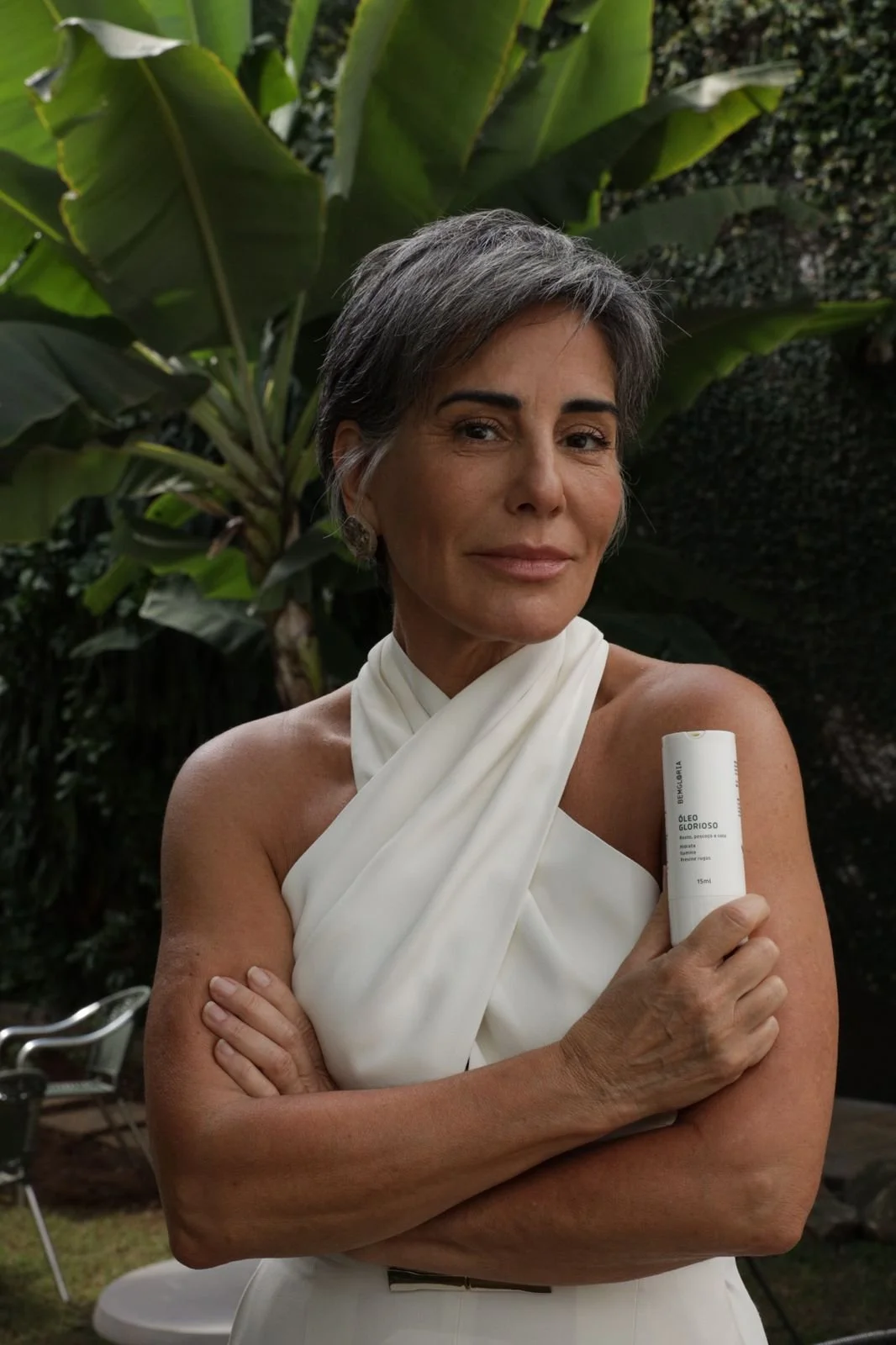 Mulher de cabelo curto e grisalho, vestindo vestido branco, segurando um tubo de óleo em um ambiente com plantas verdes ao fundo.