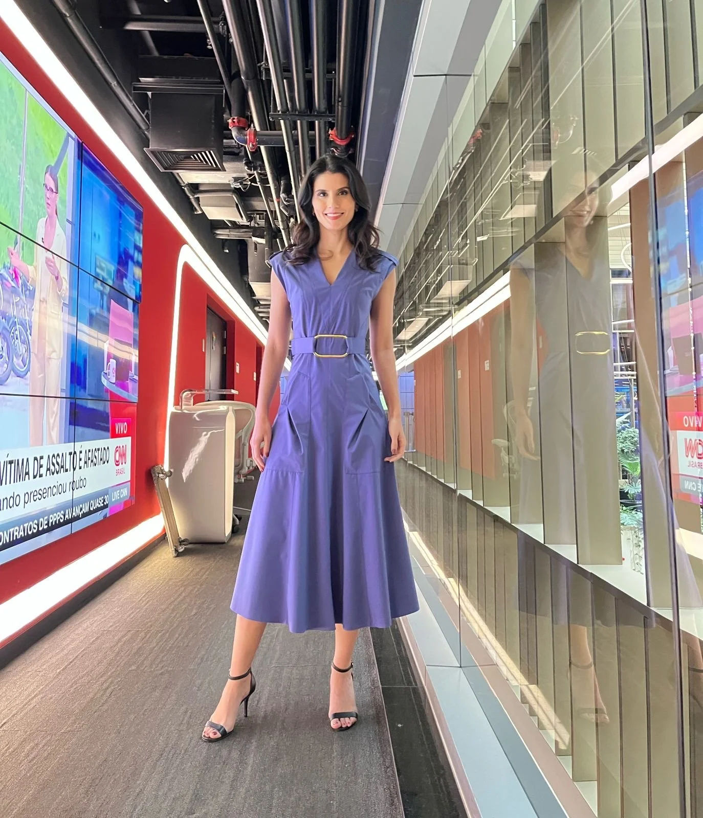 Mulher com vestido longo azul e salto alto, posando em uma televisão interativa de um jornal na sala de um aeroporto ou estação de trem.
