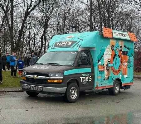 TOMS eco-friendly shoe delivery truck with colorful branding and illustrations, parked outdoors with people in the background.