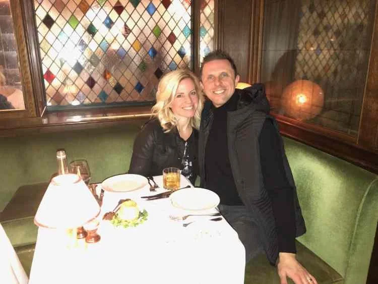 A smiling man and woman sitting close together at a restaurant table with empty plates, glasses, and a lantern, in a cozy dining setting with stained glass window and green upholstered seating.