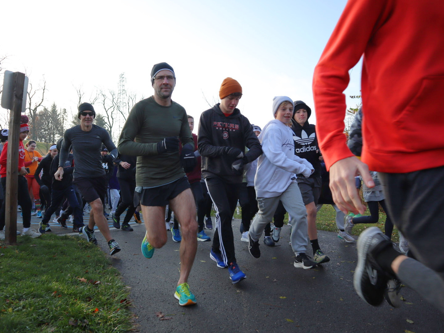 People running in a race, wearing athletic clothing and beanies, on a paved path in a park-like setting.