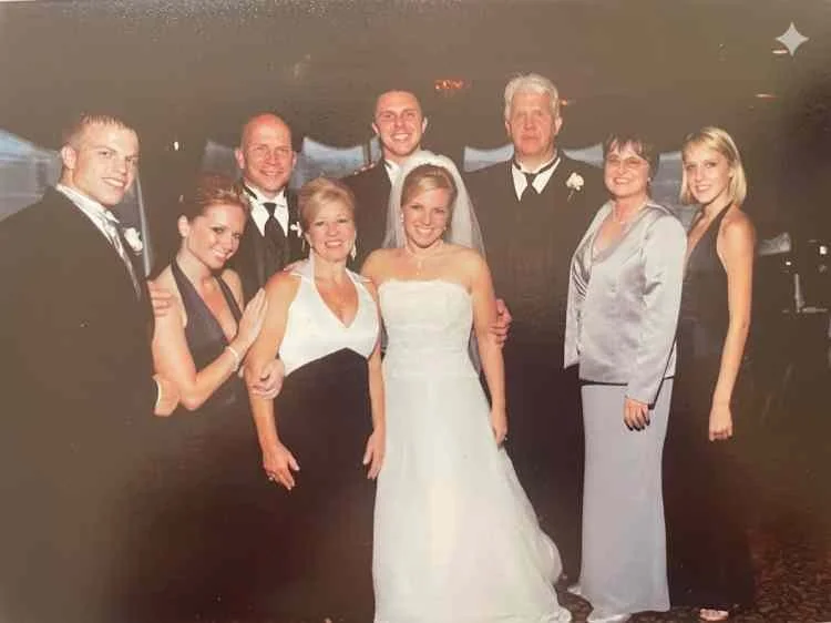 Group of nine people dressed in formal attire at a wedding celebration, with two women in wedding dresses and the rest in suits and dresses.