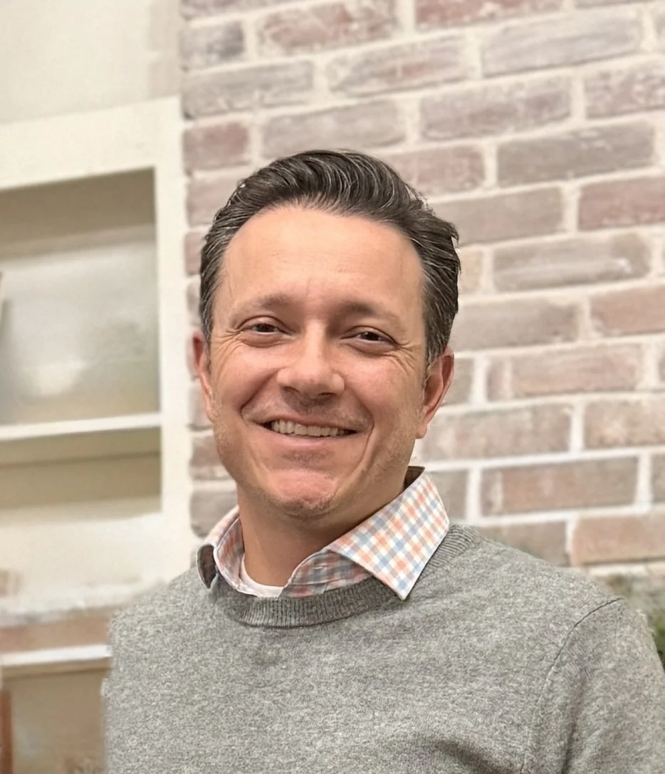 A smiling man with dark hair and a plaid shirt under a gray sweater, standing in front of a brick wall.