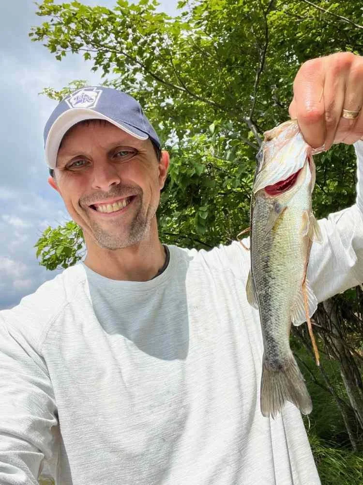Smiling man wearing a baseball cap holding a large fish outdoors in front of green trees.