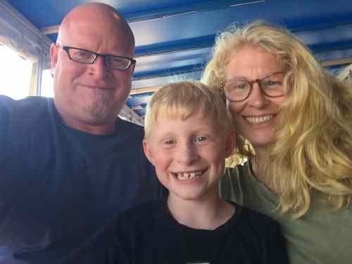 A family of three smiling together indoors, with blue walls and windows in the background.