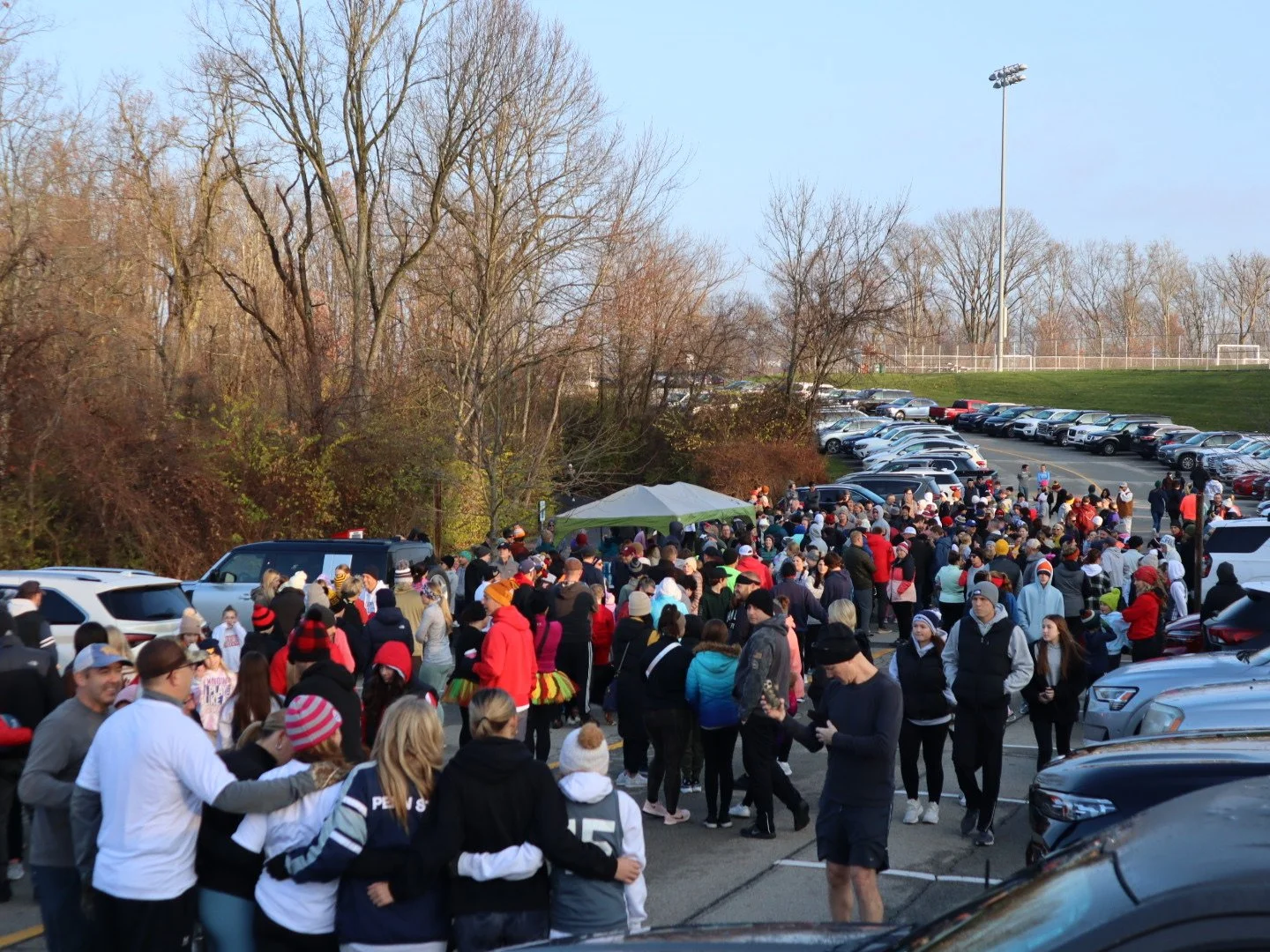 A large outdoor gathering of people in a parking lot next to a field, with some people in colorful outfits and others in casual clothing, under a clear sky with leafless trees in the background.