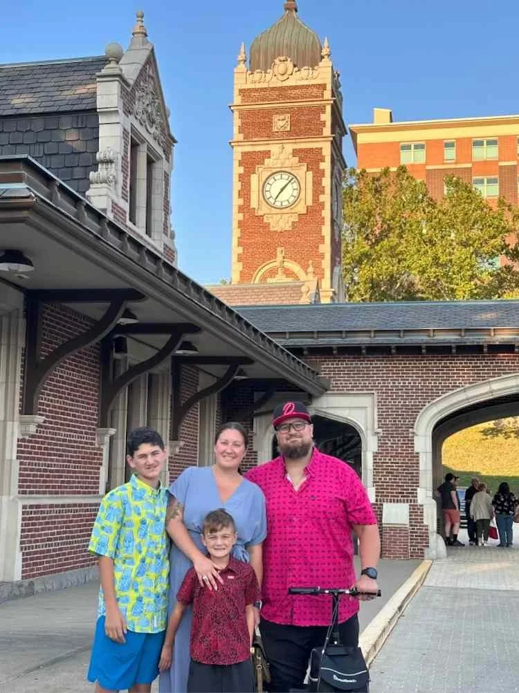 A family of four standing outside a historic train station with a clock tower in the background. The family includes a woman, a man with a scooter, and two boys, all smiling.
