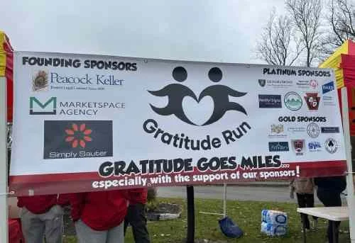 A banner for the Gratitude Run event showcasing sponsors, including Peacock Keller, Marketplace Agency, and Simply Salted. The banner features two stylized figures holding hands, with the text 'Gratitude Goes Miles' and 'especially with the support of our sponsors!' at the bottom.