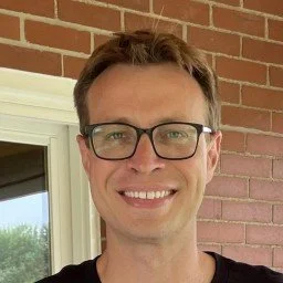 A man with short brown hair wearing glasses and a black shirt, smiling in front of a brick wall and window.