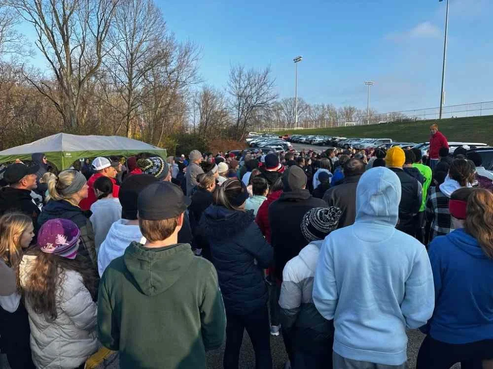 Crowd of people gathered outdoors near a parking lot, some wearing jackets and hats, with a man speaking to the crowd.