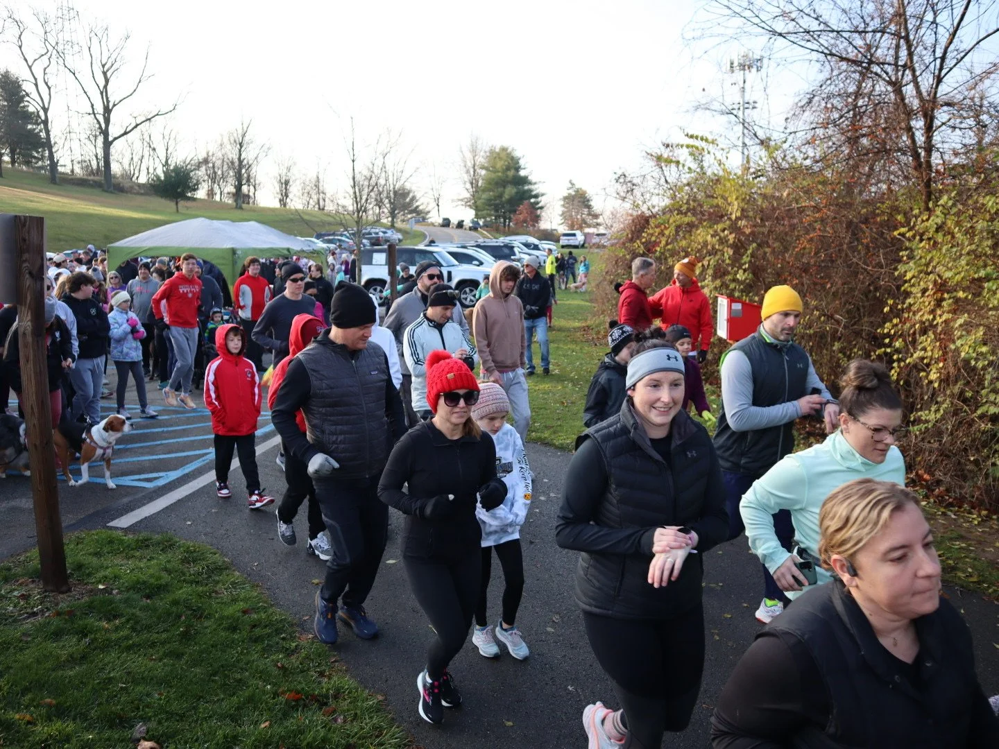 People participating in an outdoor run or walk event on a fall day, with some wearing athletic gear and others in casual clothing, trees with autumn leaves, and a parking area in the background.