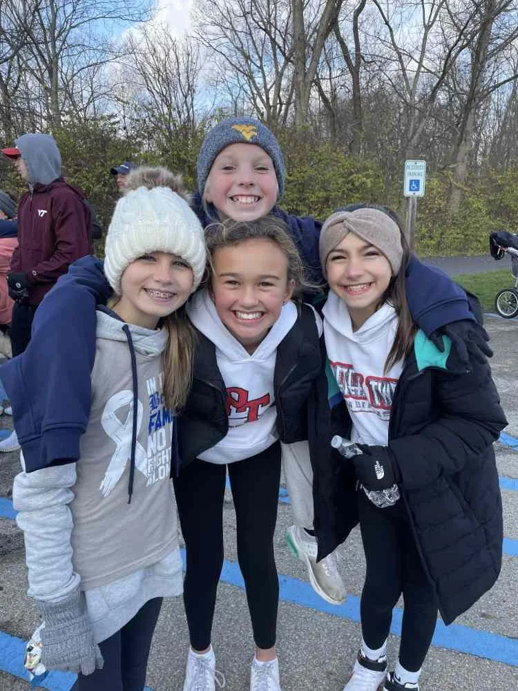 Four young girls smiling and hugging outdoors, dressed warmly in jackets and hats, with trees in the background.