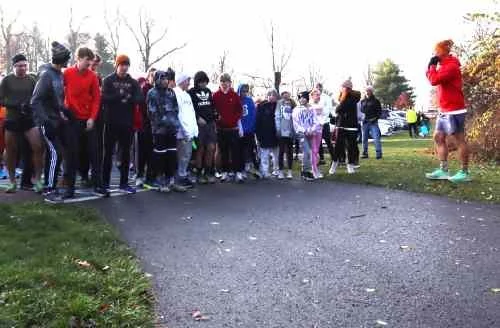 A group of people, including children and adults, standing in a line outdoors on a paved path, with a woman in red speaking to them. Trees and cars are visible in the background.