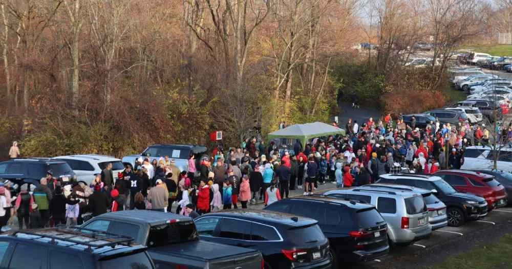 A large crowd of people standing in line outdoors next to parked cars in a wooded area during autumn.