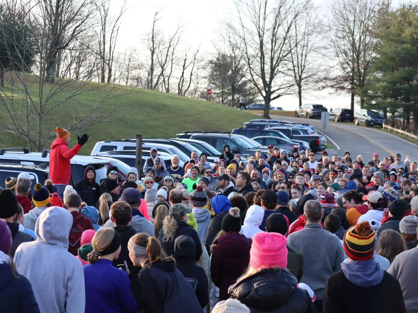 A large crowd gathered outdoors for a rally or event, with a person speaking to the crowd while wearing a red hoodie and brown hat, and cars parked in the background on a grassy area with trees.