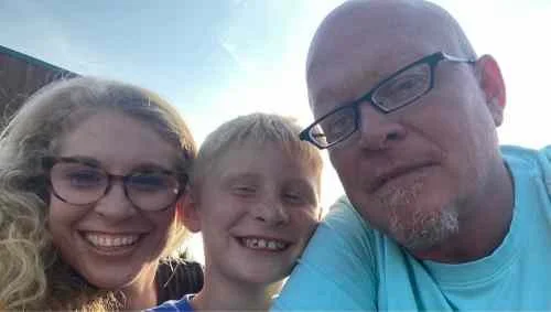 A woman, a young boy, and an older man with glasses smiling outdoors on a sunny day.