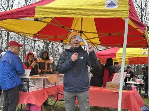 A group of people at an outdoor event under yellow and red tents, with a woman speaking into a microphone in the foreground.