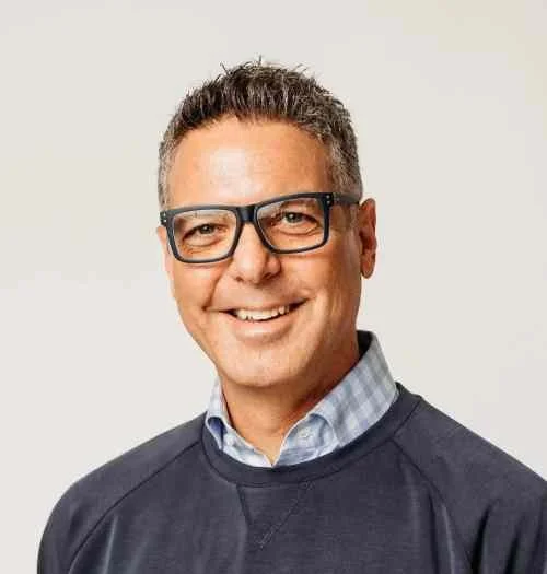 A man with short dark hair, glasses, and a light blue collared shirt smiling and wearing a dark sweatshirt against a plain background.
