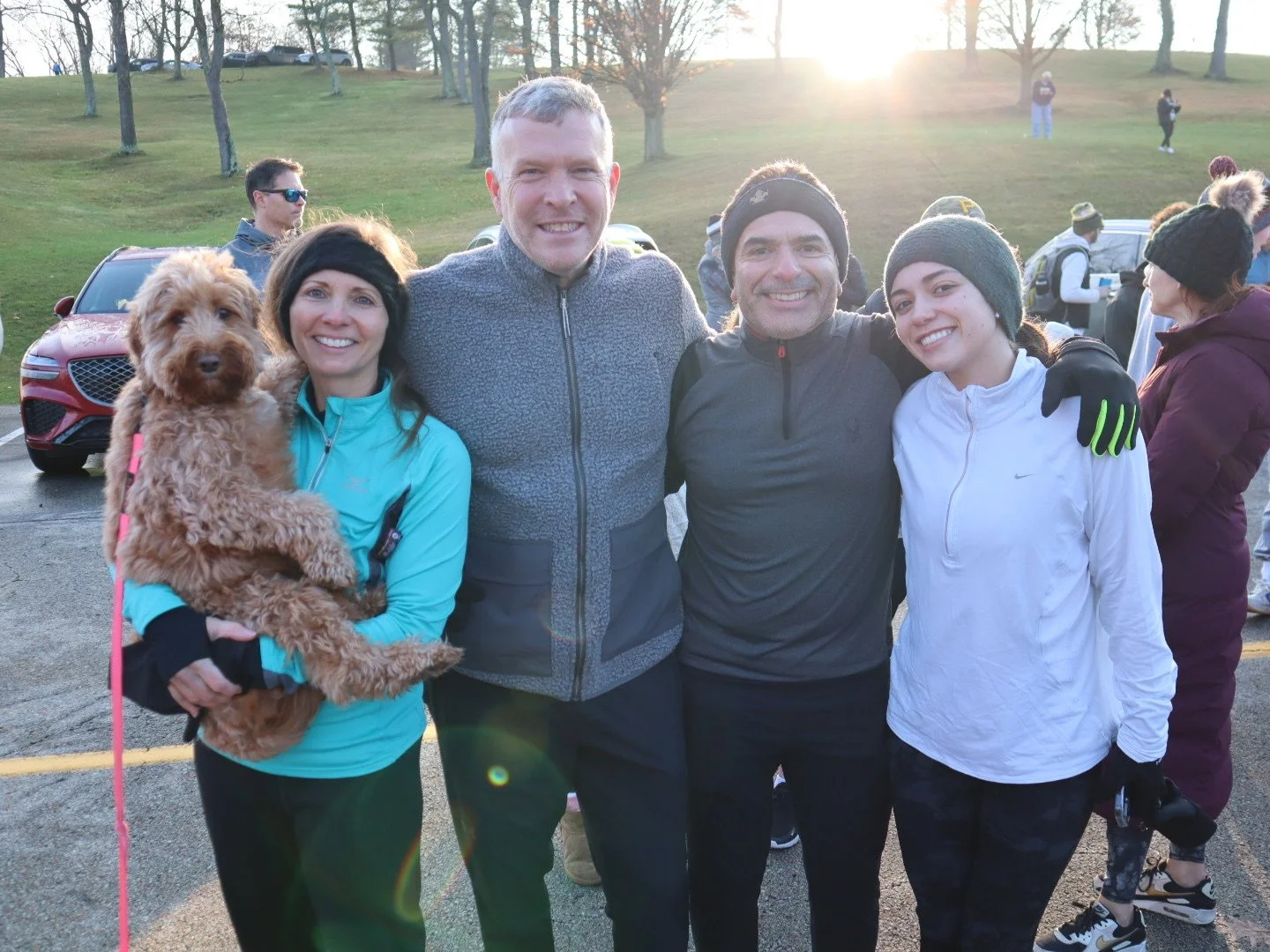 Four people smiling and standing close together outdoors at a park, with one woman holding a brown dog. The sun is setting behind trees in the background, and other people are visible in the distance.