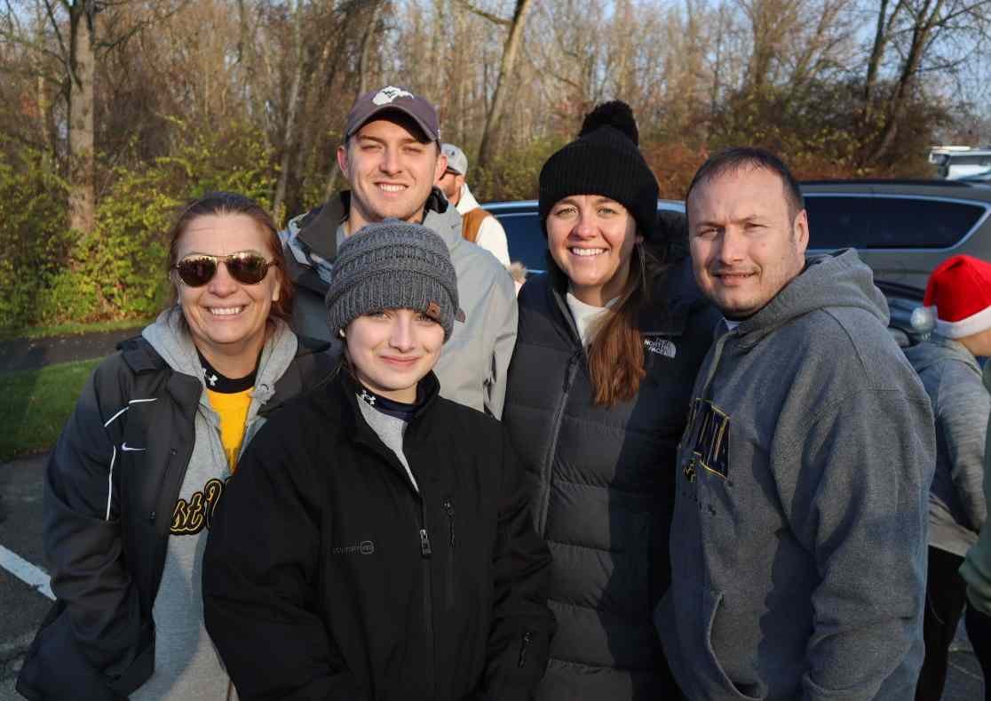 Group of five people outdoors in a parking lot, smiling, wearing jackets and hats, with trees and parked cars in the background, some wearing holiday accessories.