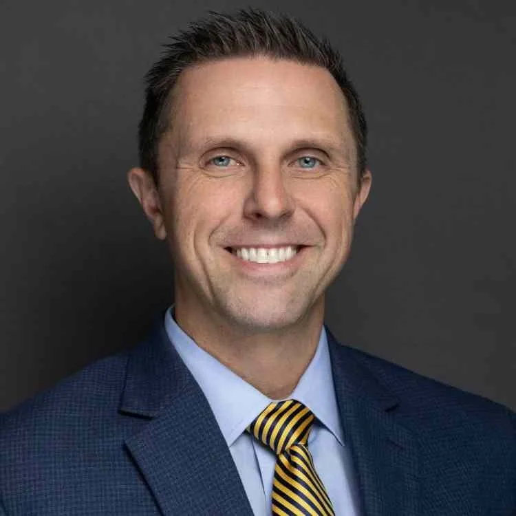 Professional headshot of a smiling man in a navy suit, light blue shirt, and yellow striped tie, against a dark gray background.